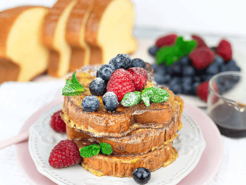 Three slices of French toast stacked on a plate, topped with powdered sugar, fresh berries, and mint, with a side of syrup and sliced bread in the background.