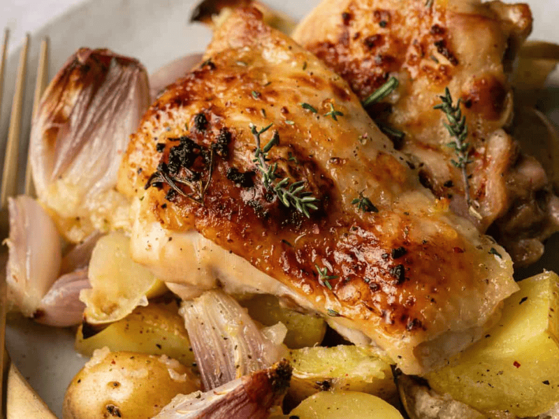 Roast chicken thighs with crispy skin served on a plate with roasted potatoes, onions, and fresh herbs, next to a gold-colored fork and knife.
