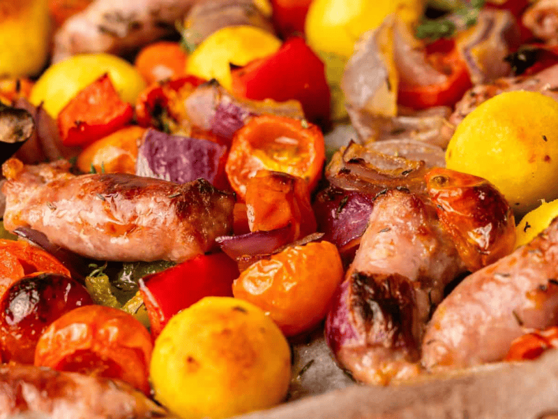 Roasted sausages, potatoes, cherry tomatoes, red onions, and bell peppers mixed together on a baking tray.