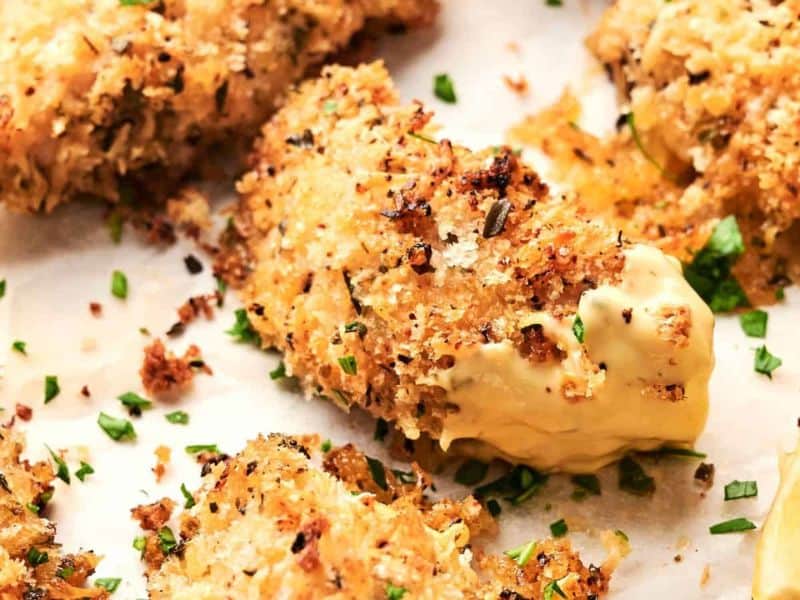 Close-up of crispy, Sheet Pan Parmesan Crusted Chicken pieces garnished with chopped herbs and served with a lemon wedge. The pieces are placed on a parchment-lined surface.