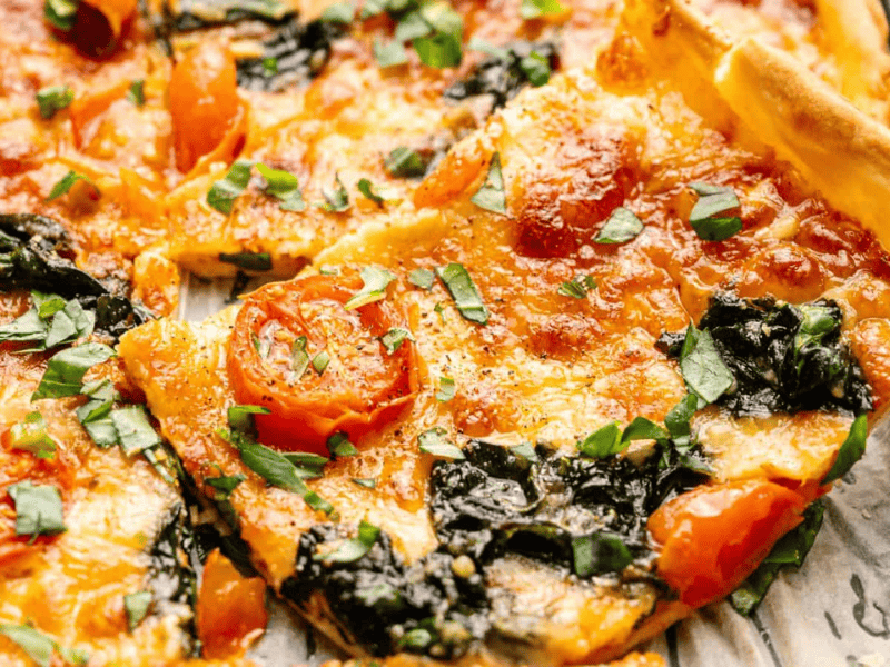 A close-up of a rectangular pizza topped with melted cheese, cherry tomatoes, spinach, and fresh herbs, on a baking tray lined with parchment paper.