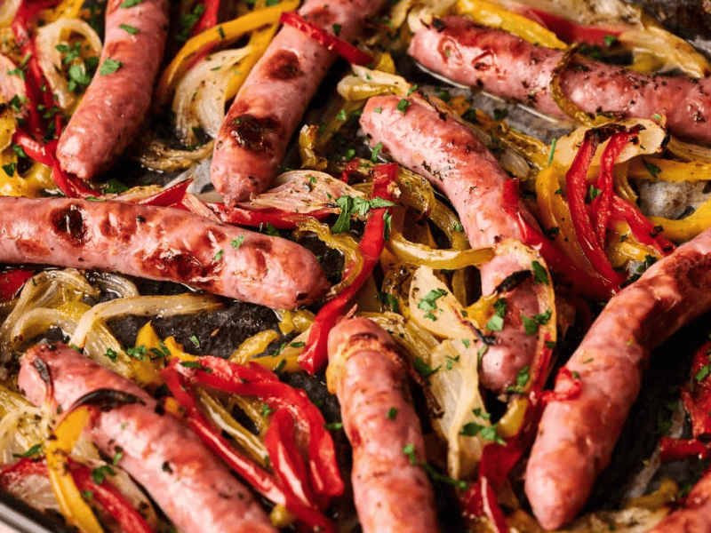 Sheet pan with roasted sausages, sliced red and yellow bell peppers, and onions, garnished with chopped parsley.