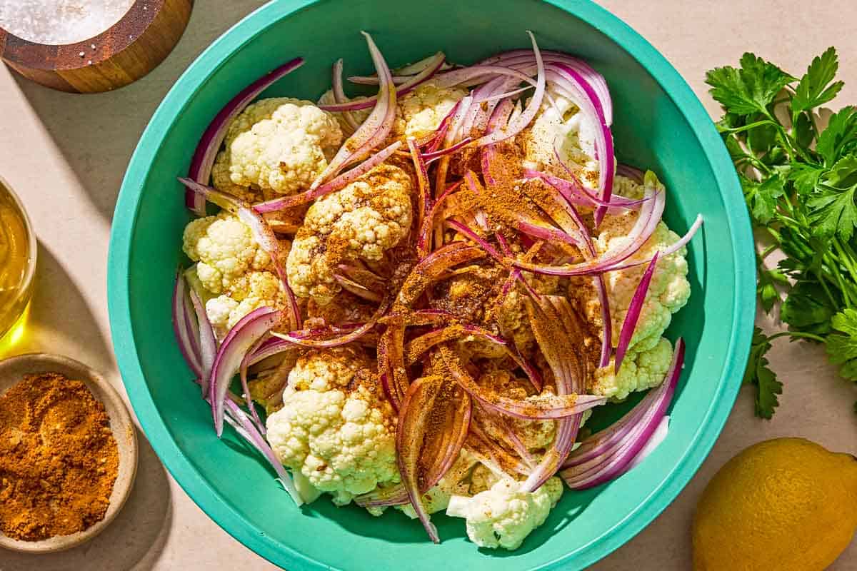 The ingredients for the air fryer cauliflower in a bowl just before being mixed together. next to this is a lemon, parsley, a bowl with ras al hanout, a bowl of salt, and a bottle of olive oil.