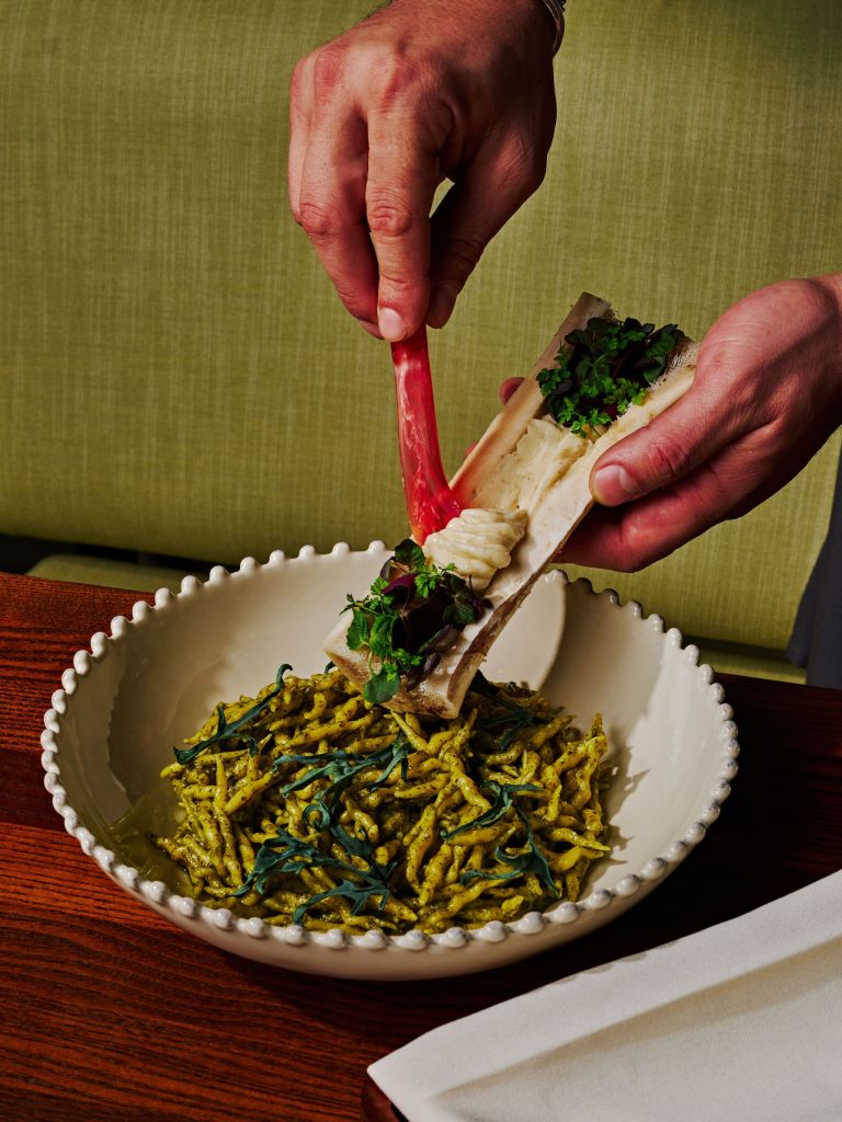 Chef plates bone marrow over pesto trofie pasta with fresh herbs in a scalloped white bowl at a local restaurant.