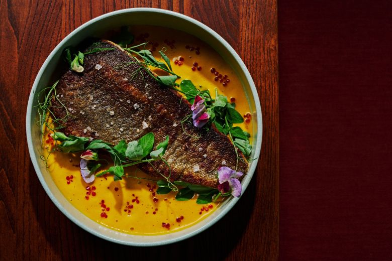 Crispy-skin fish fillet over golden sauce, garnished with pea shoots, edible flowers, and chili oil in a ceramic bowl.