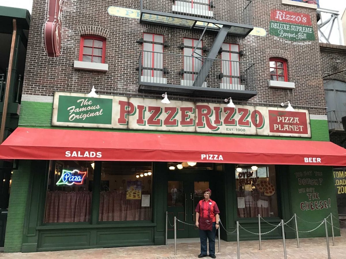A person stands outside PizzeRizzo in Muppets Courtyard, a pizza spot with a red awning and brick exterior.