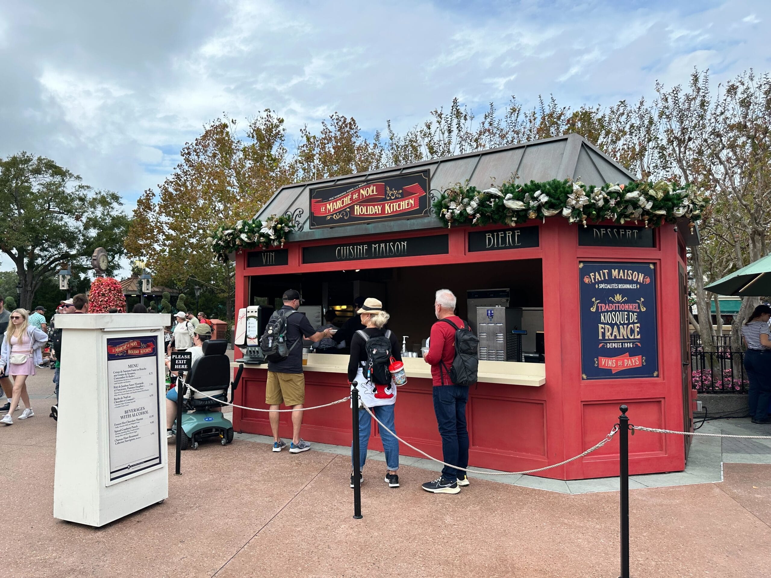 People gather around a festive outdoor food kiosk adorned with Christmas decorations at the EPCOT International Festival of the Holidays. Signs and a menu board invite visitors to explore delightful seasonal treats.