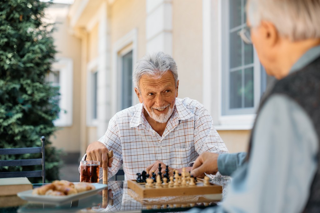 Two senior men playing chess and talking on a patio.