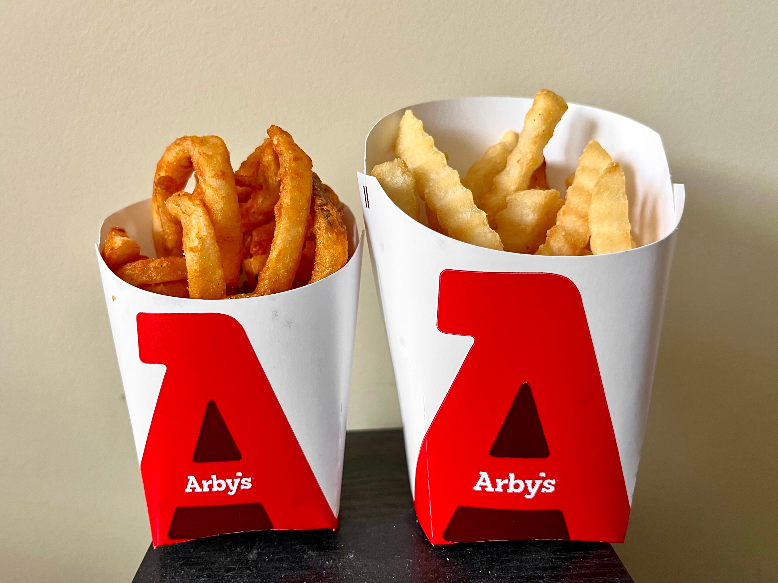 Two Arby’s containers, one filled with crispy curly fries and the other with crinkle-cut fries, are placed side by side against a beige background.