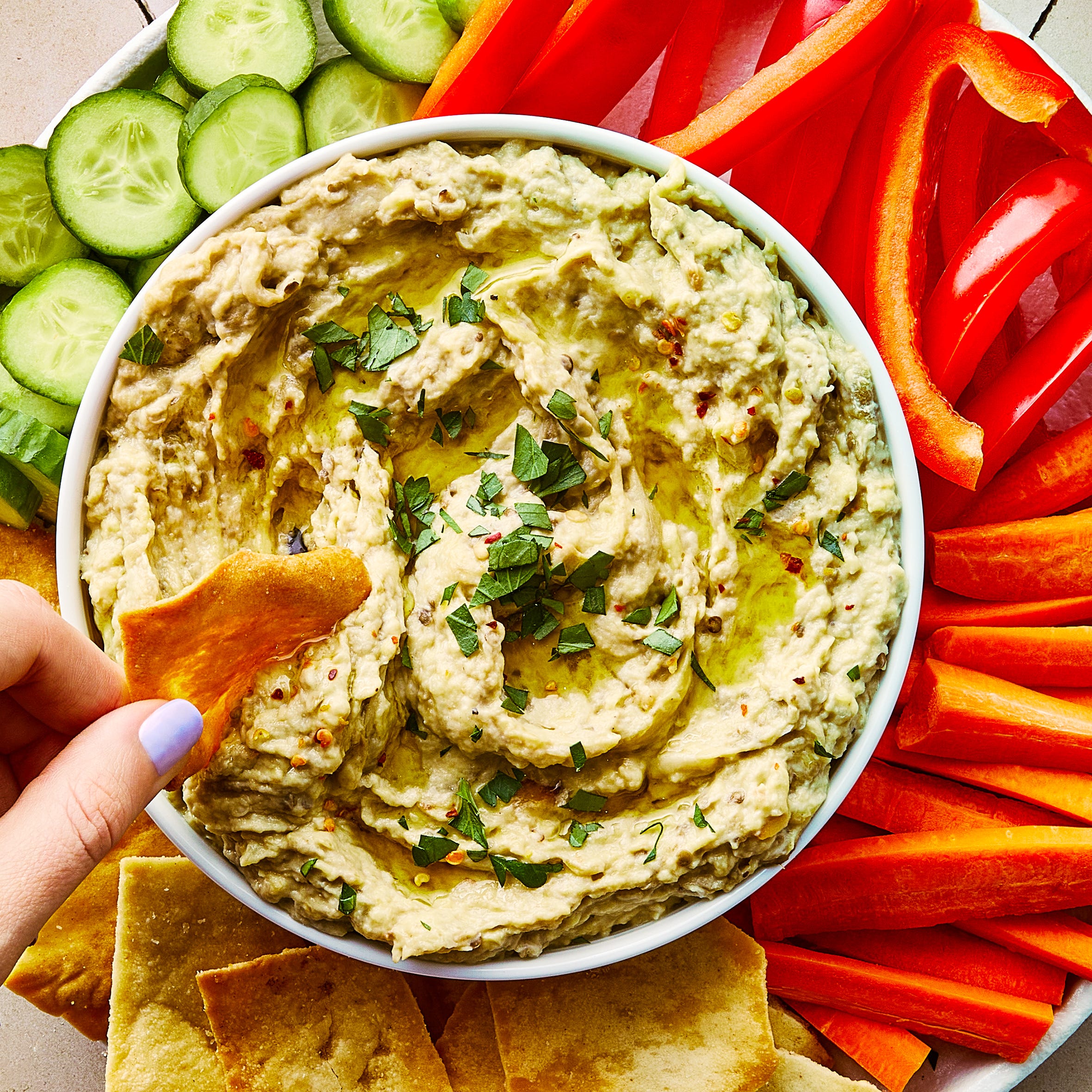 baba ganoush in a bowl topped with red pepper flakes and chopped parsley, and surrounded by pita chips and crudite