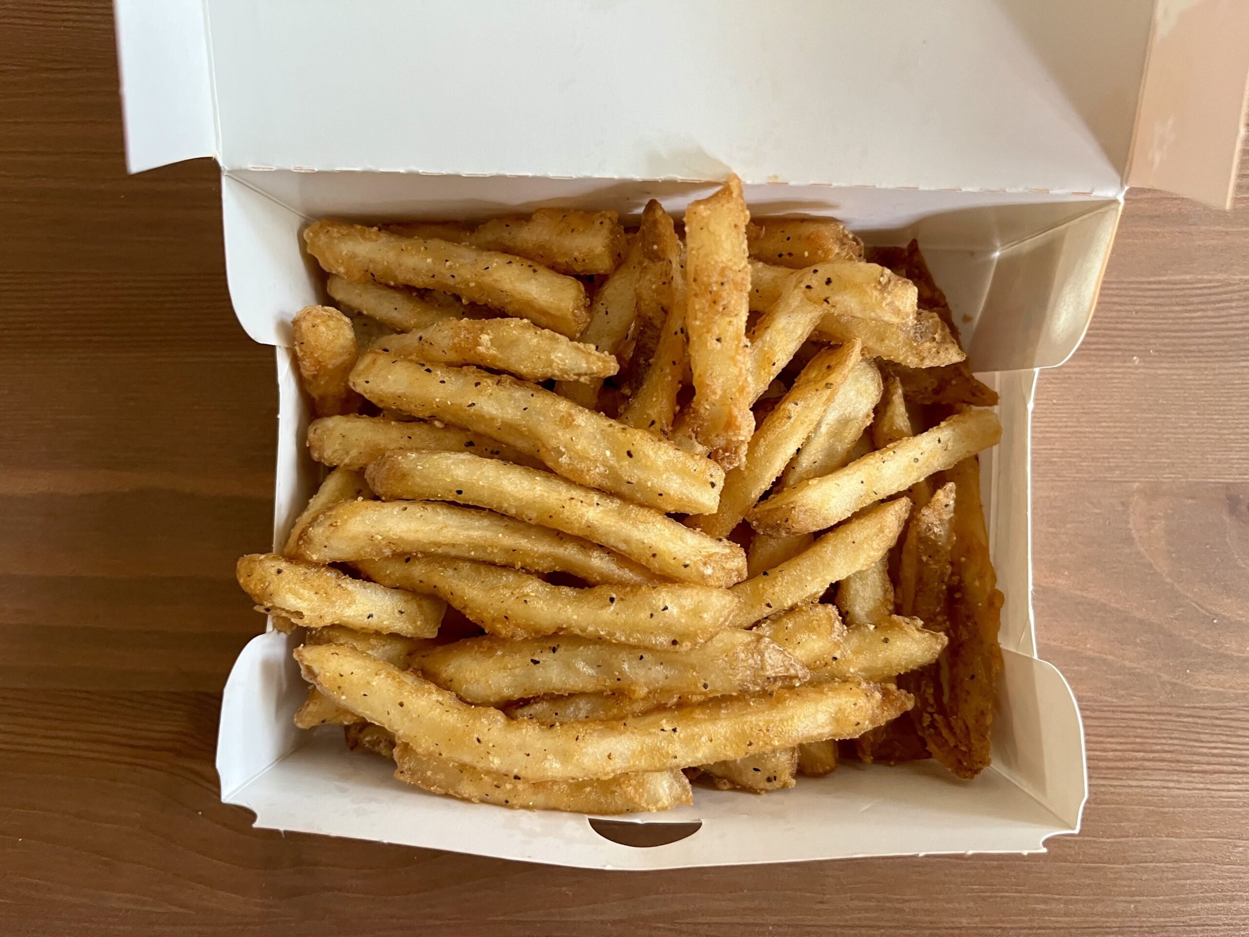 A white takeout box filled with seasoned French fries sits on a wooden surface. The fries are golden brown and sprinkled with visible black pepper and spices.