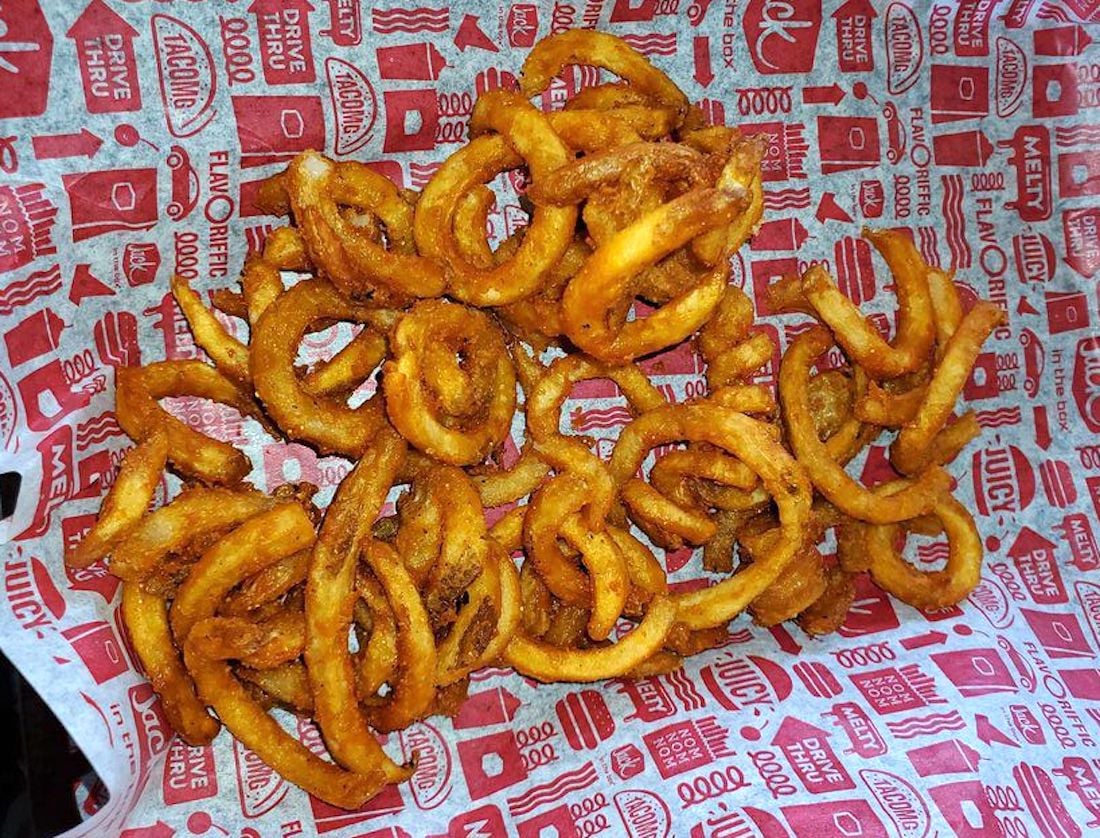A serving of seasoned curly fries is spread over a red and white paper liner with repeating fast food-themed text and icons. The fries are crispy and golden brown.