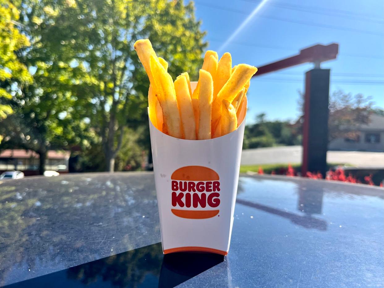 A Burger King container of French fries sits on a shiny surface outdoors, with trees and a sunny sky in the background.