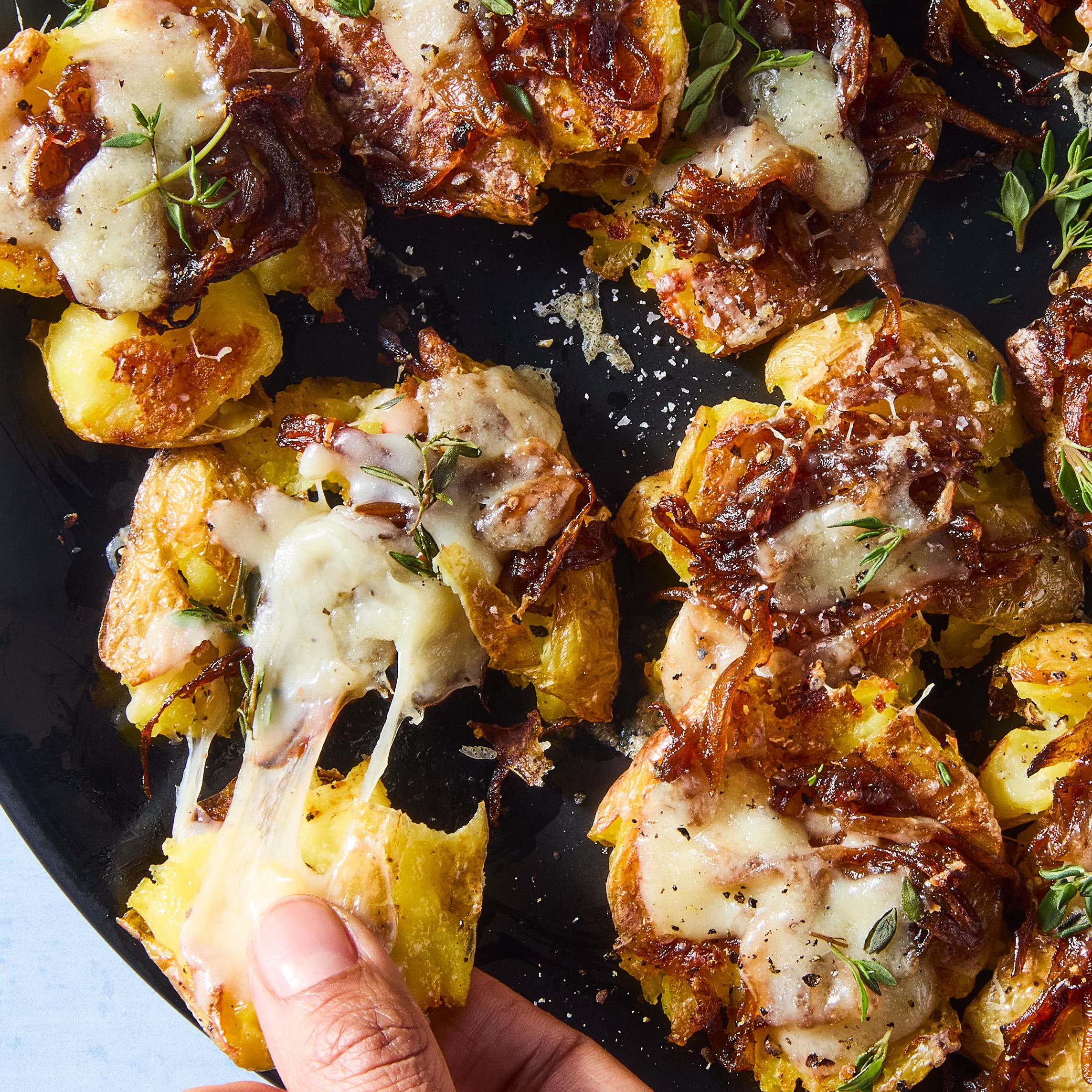 french onion smashed potatoes topped with caramelized onions, gruyere, and thyme on a black plate