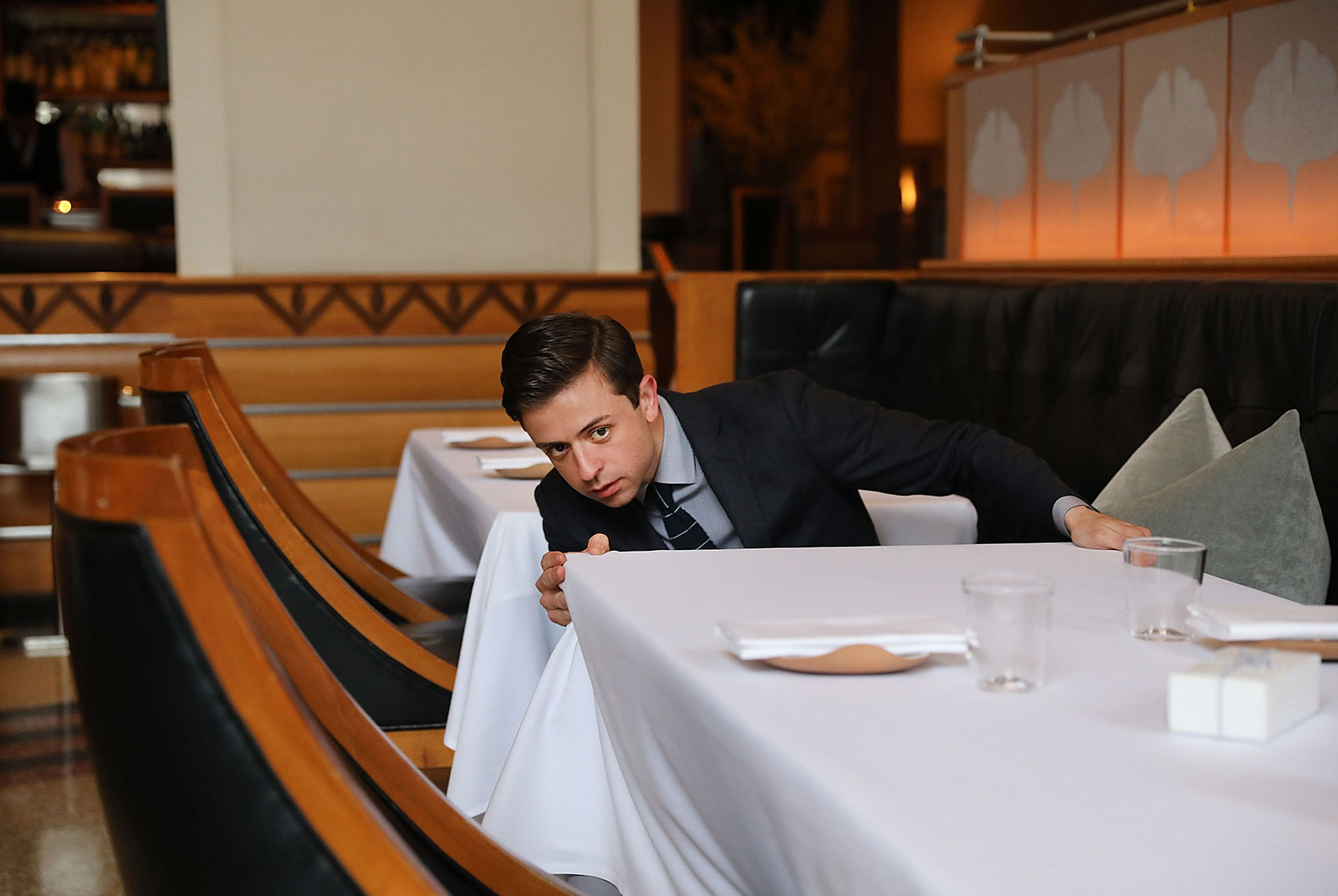 Person in suit kneeling beside table, carefully inspecting tablecloth at eye-level
