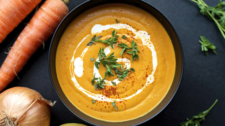 A bowl of carrot and onion soup next to herbs and veggies