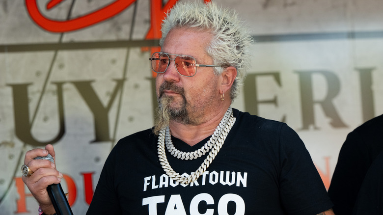 A spiky, blonde-haired Guy Fieri holds a mic, wearing shades and a chunky chain necklace