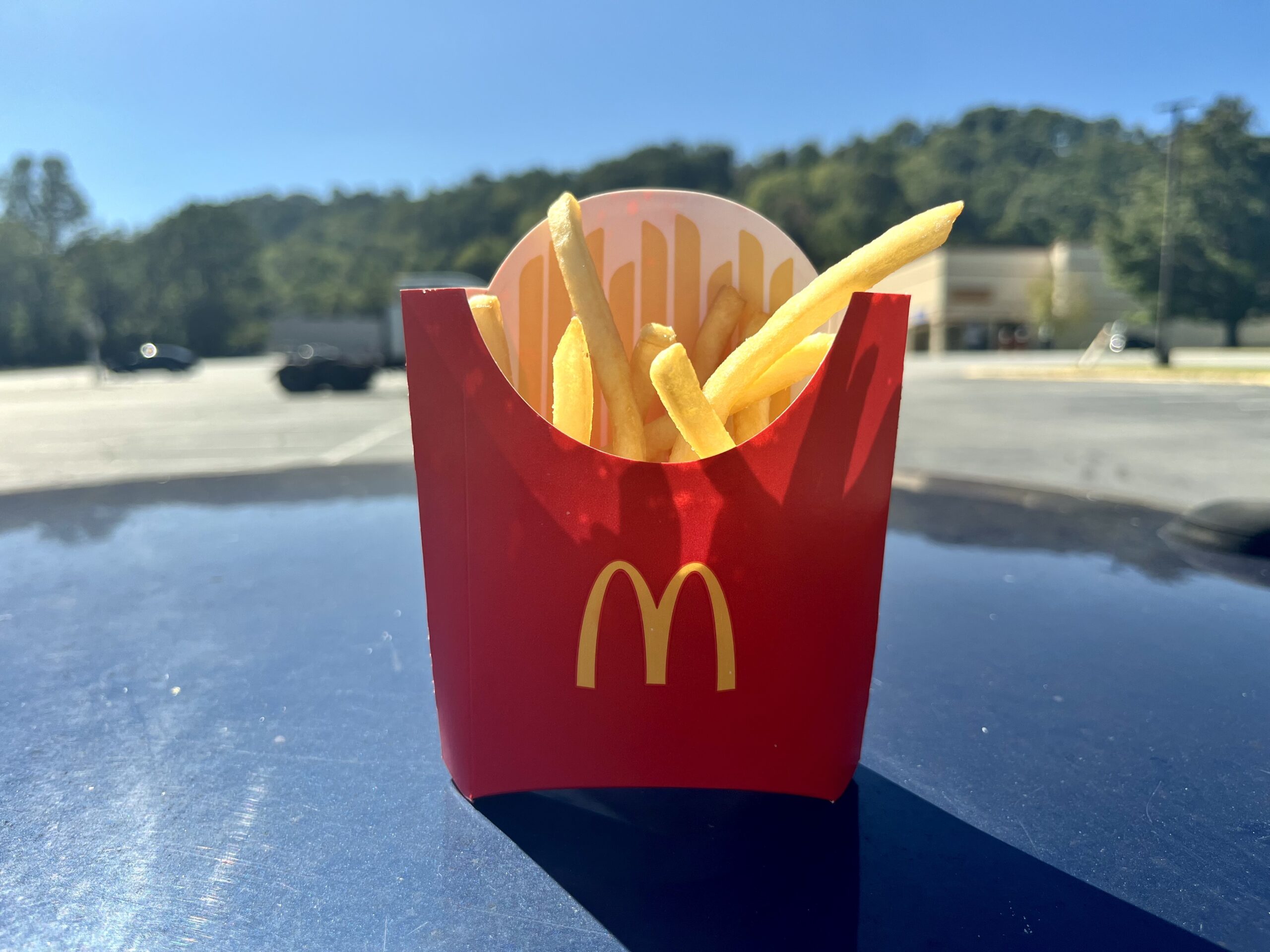 A red McDonald's container filled with French fries sits on a car hood in a sunny parking lot, with trees and buildings blurred in the background.