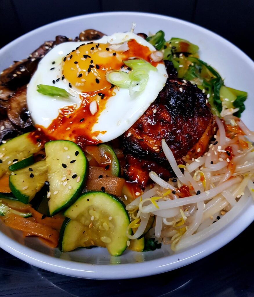 Bibimbap - Korean Rice Bowl. Gochujang Chicken, Sweet Soy Mushrooms, Blanched and Marinated Pak Choi, Beansprouts, Carrots and Courgettes. Finished with an Egg, Seeds and Sauce.