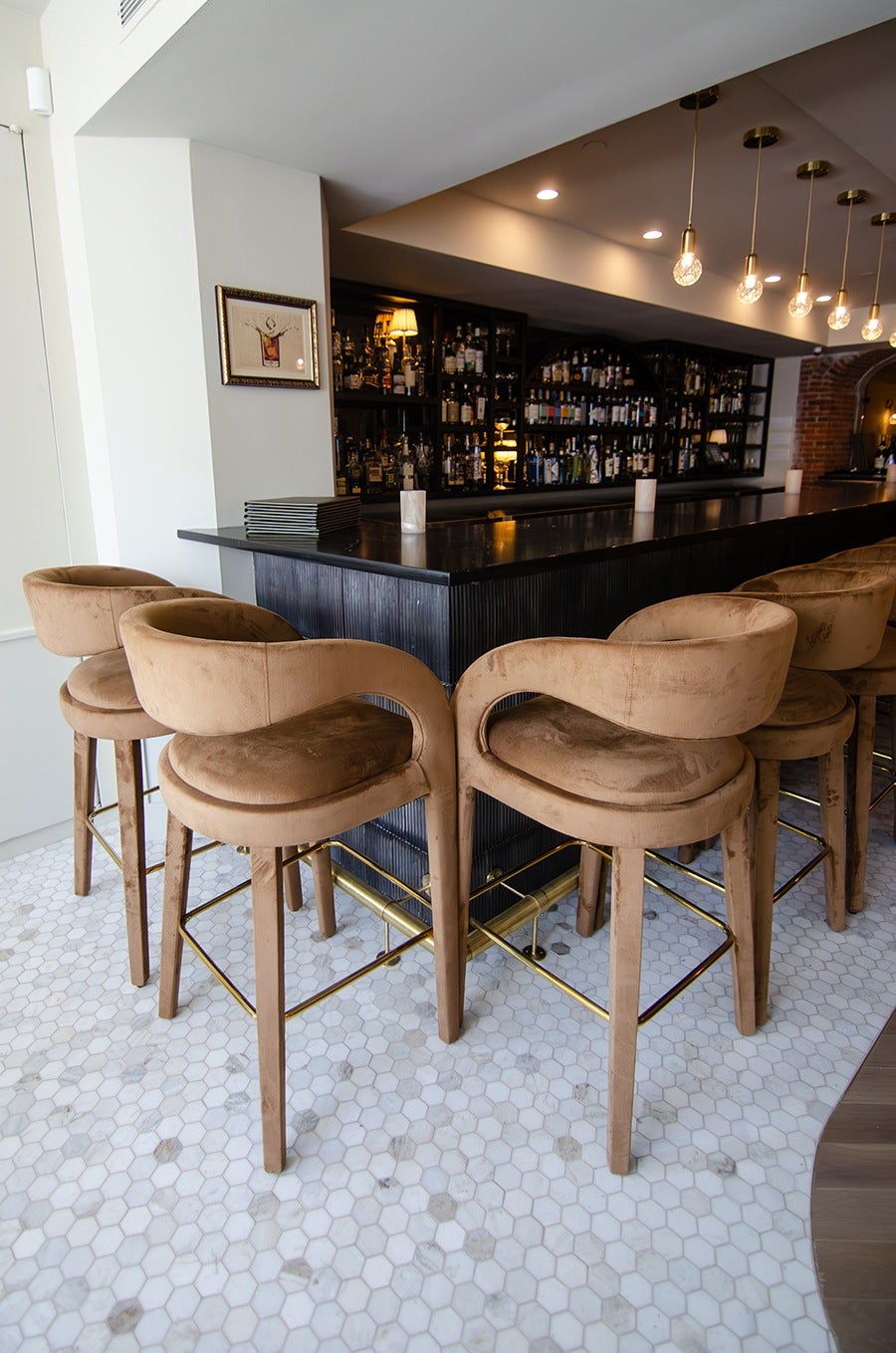 A bar area at a restaurant features light brown velvet high chairs at a dark wooden bar on a white tiled floor.
