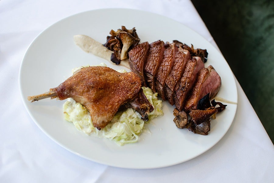 An elegantly plated dish of duck two ways, including seared and sliced breast and confit leg, with mushrooms and other garnishes.