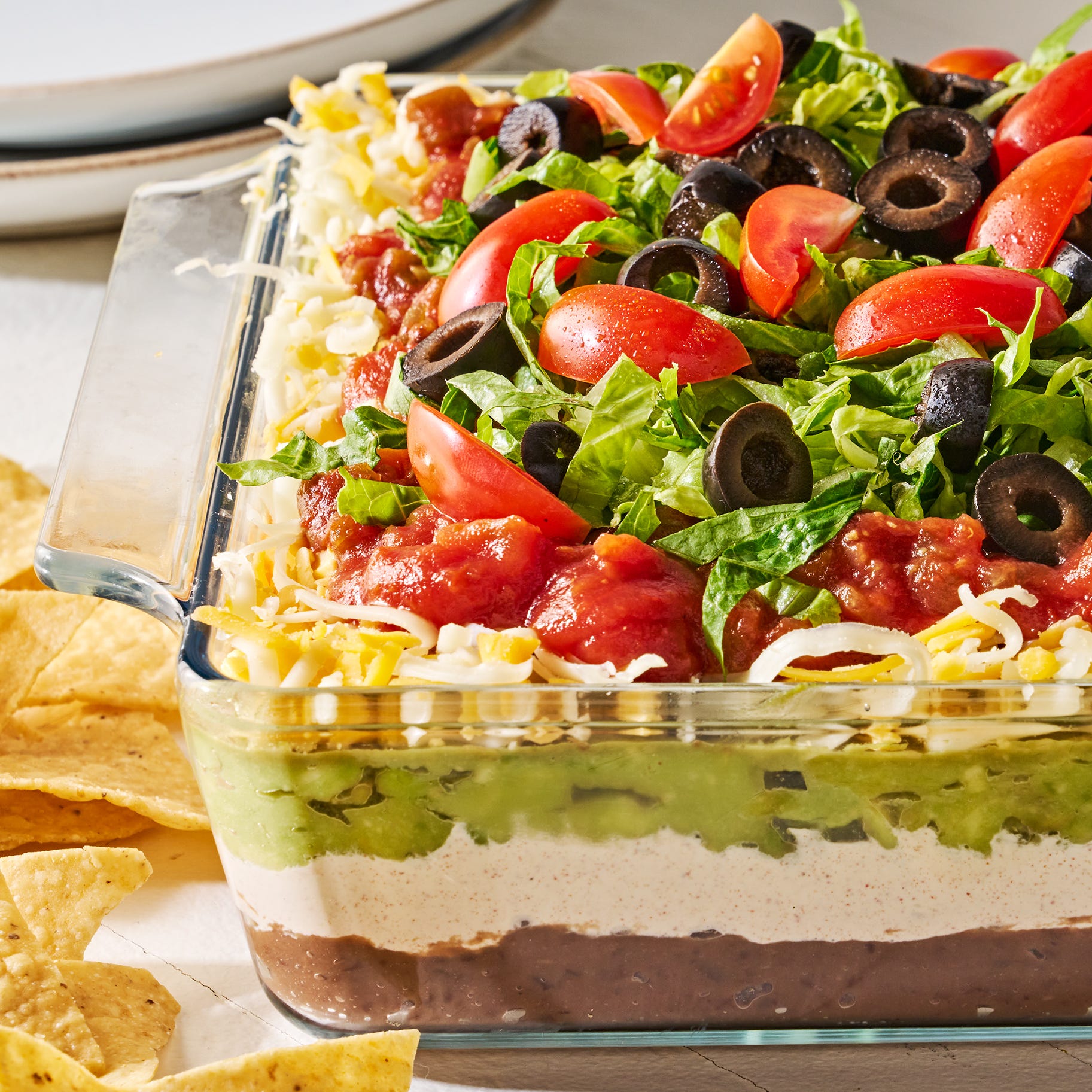 layered dip with refried beans, sour cream, guacamole, cheese, tomatoes black olives, and lettuce in a clear dish