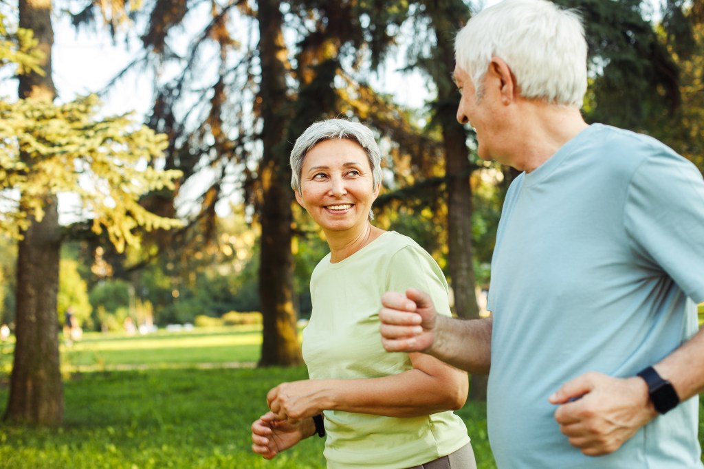 A senior man and woman jogging outdoors, looking at each other and smiling.