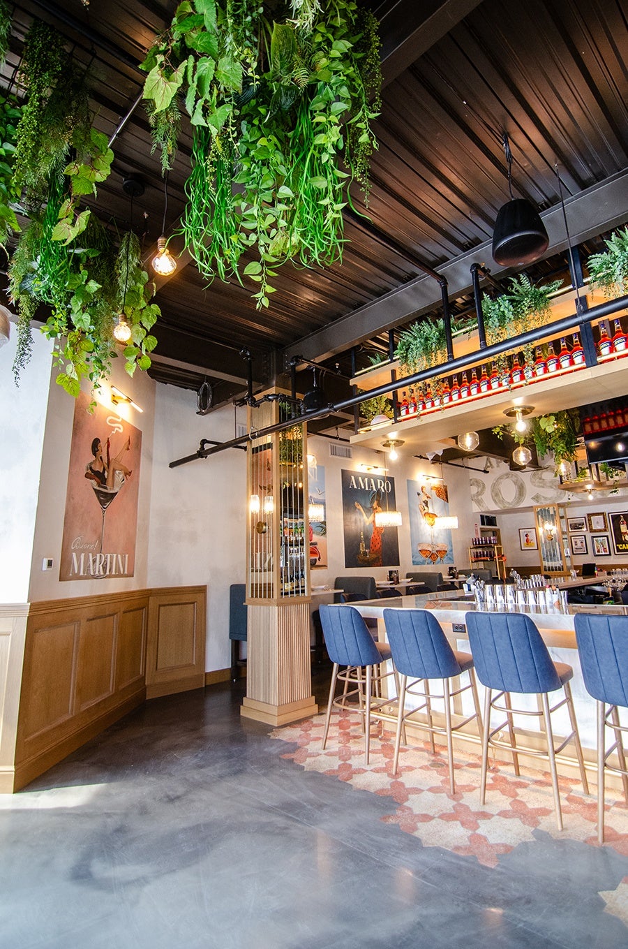 Modern bar interior with a polished concrete floor and patterned tile near the bar. The bar features a white marble countertop with blue upholstered bar stools with gold legs. Above the bar, there is a suspended shelf holding bottles and greenery. The ceiling is dark with hanging plants and exposed industrial lighting. The walls have framed posters and artwork, including a large 