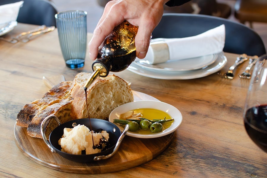A hand is pouring balsamic vinegar from a dark bottle onto a small black dish containing cheese. The dish is on a wooden board alongside slices of rustic bread and a small white plate with green olives, garlic cloves, and rosemary in olive oil. The setting includes a wooden table with a blue glass, white plates with a folded napkin, and silver cutlery.