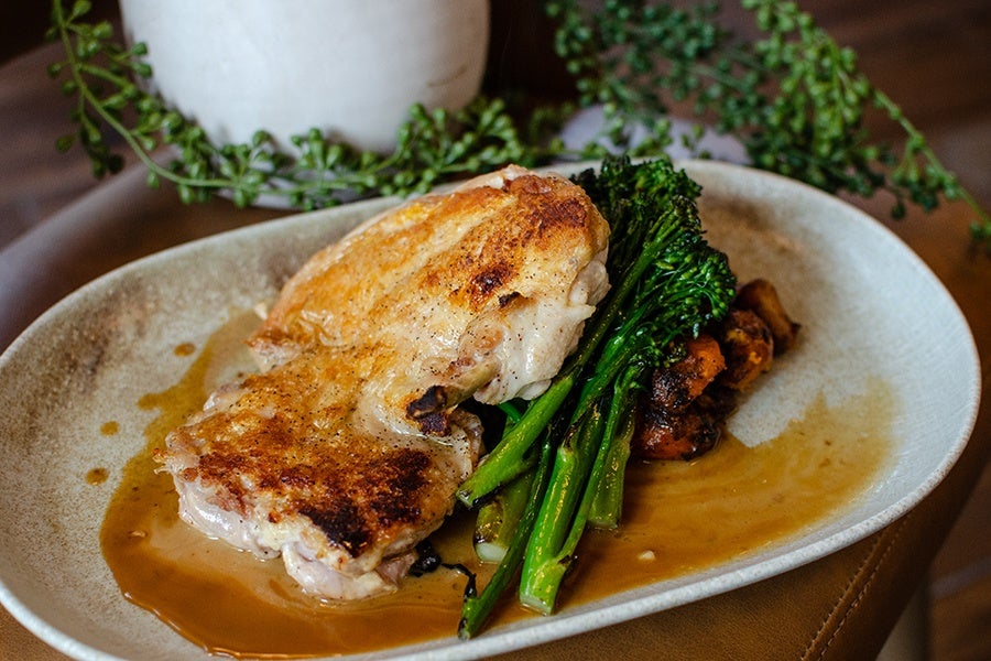 A plate with a serving of roasted chicken accompanied by sautéed broccolini and roasted carrots, all drizzled with a light brown sauce. The dish is presented on a beige, oval ceramic plate with a rustic background featuring green herbs.