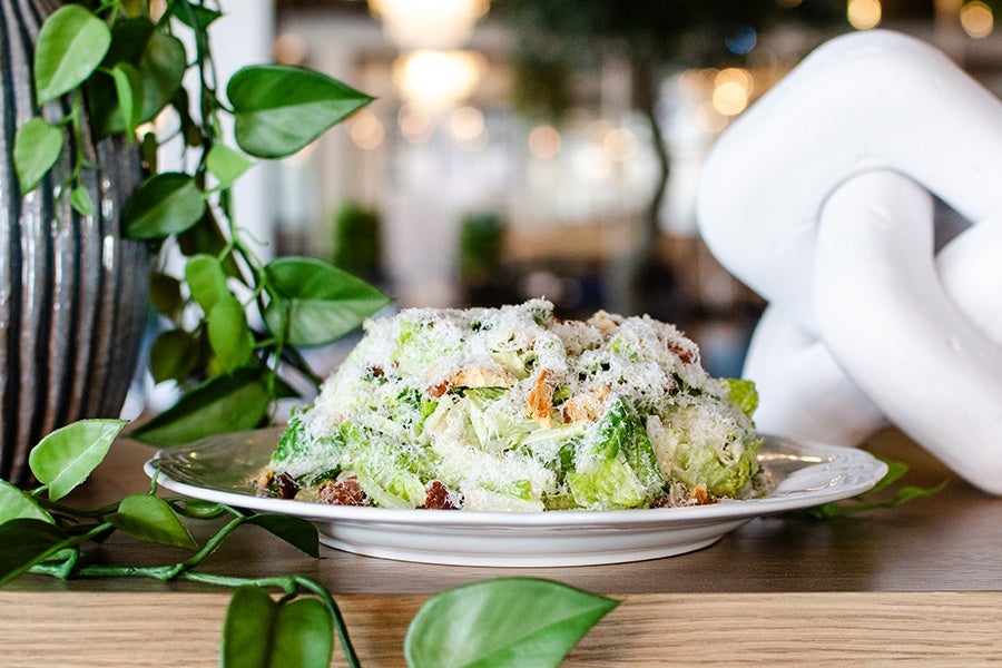 A plate of Caesar salad topped with grated cheese, placed on a wooden surface. Green leafy plants and a large white decorative object are visible in the background.