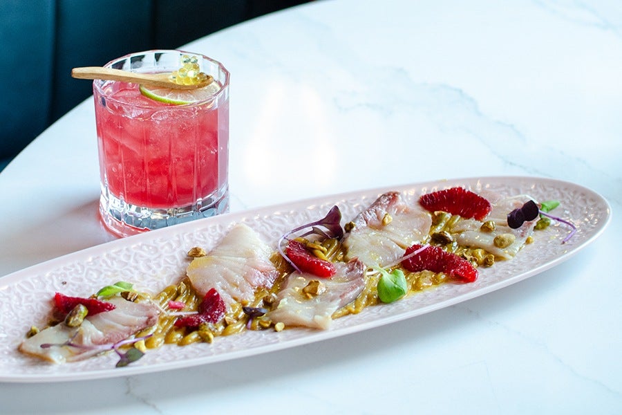 A long, oval pink plate with thin slices of raw fish garnished with red citrus segments, microgreens, and crushed nuts, accompanied by a pink cocktail in a clear glass with ice, a lime slice, and a wooden spoon topped with small yellow spheres. The setting is on a white marble table.