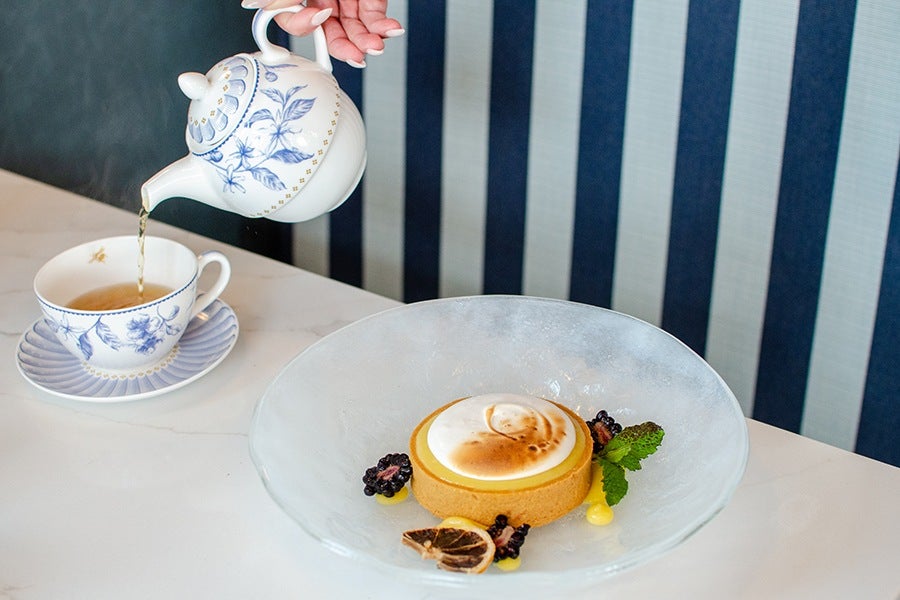 A lemon tart with toasted meringue on top, garnished with blackberries, a dried lemon slice, mint leaves, and small dollops of yellow sauce, served on a clear glass plate. Next to it, tea is being poured from a white teapot with blue floral designs into a matching teacup and saucer. The background features a blue and white striped wall.