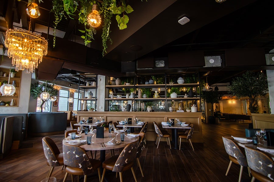 Modern restaurant interior with wooden floors, round and rectangular tables set with glassware and napkins, patterned cushioned chairs, hanging plants, and warm ambient lighting from chandeliers and bulbs. Shelves with decorative vases and greenery divide the space.