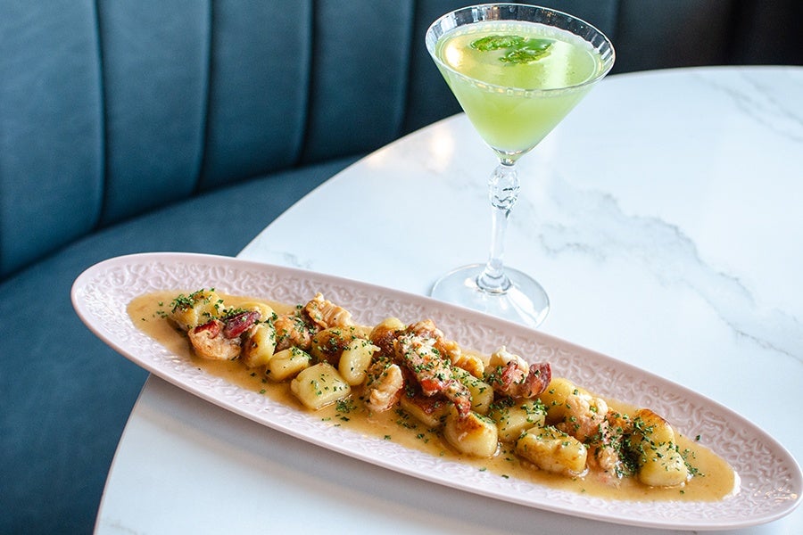 A long, oval pink plate holds a serving of gnocchi in a creamy sauce, garnished with herbs and pieces of lobster or crab. Next to the plate is a clear martini glass filled with a light green cocktail, garnished with fresh mint leaves. The setting is on a white marble table with a blue cushioned bench in the background.