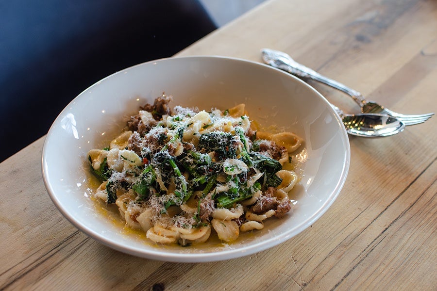A white bowl filled with orecchiette pasta mixed with cooked greens, crumbled sausage, and grated cheese, placed on a wooden table next to a fork and spoon.