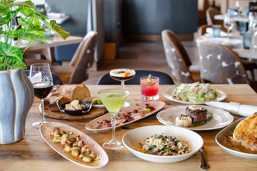 A wooden table set with a variety of dishes and drinks, including a plate of gnocchi with sauce, a bowl of pasta with greens and cheese, a steak garnished with roasted garlic and herbs, a salad, and toasted bread. There are also three cocktails: a green martini, a red drink with a garnish, and a creamy cocktail with an orange peel. A glass of red wine and a small dish of bread with butter are also present. A large vase with green leafy plants is on the left side of the table. The background shows a restaurant setting with chairs and tables.