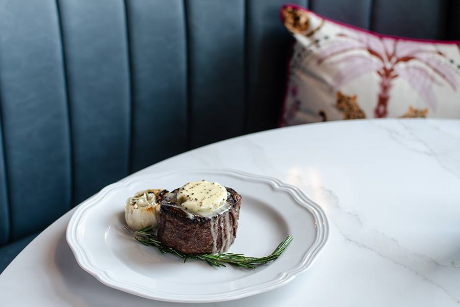 A cooked filet mignon steak topped with a pat of melting herb butter, served on a white plate with a sprig of rosemary and a roasted garlic bulb, placed on a white marble table with a blue upholstered bench and a decorative pillow in the background.