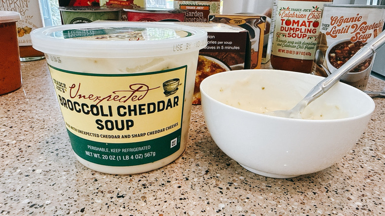 Trader Joe's Unexpected Broccoli Cheddar soup next to some in a white bowl with other TJ's soups in background