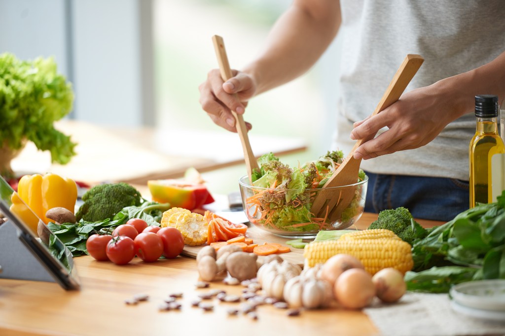Man mixing a vegetable salad with wooden utensils in a glass bowl.
