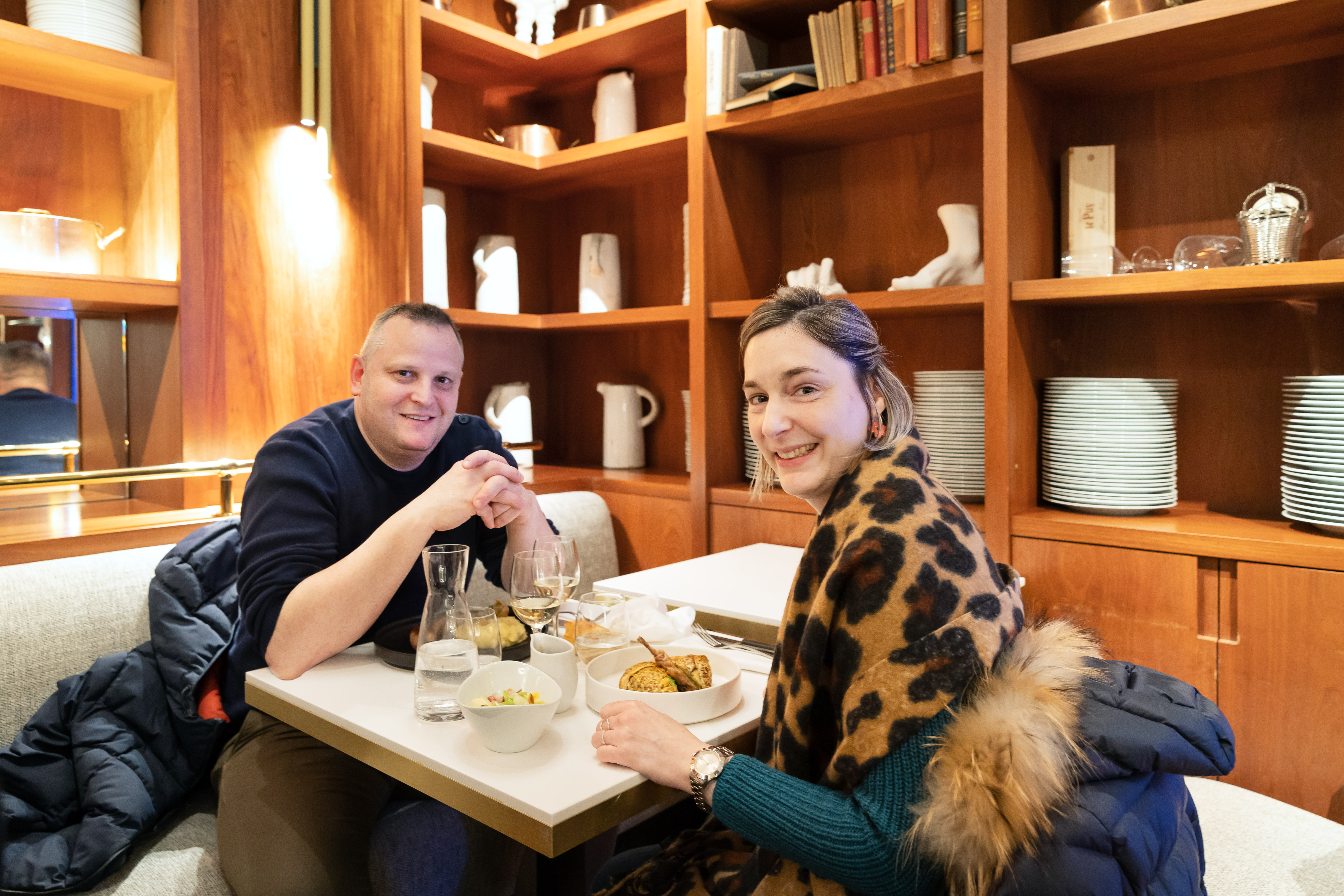 Caroline and Oliver CLOTEAU seated at a table with plates of food and glasses of wine, looking at the camera.