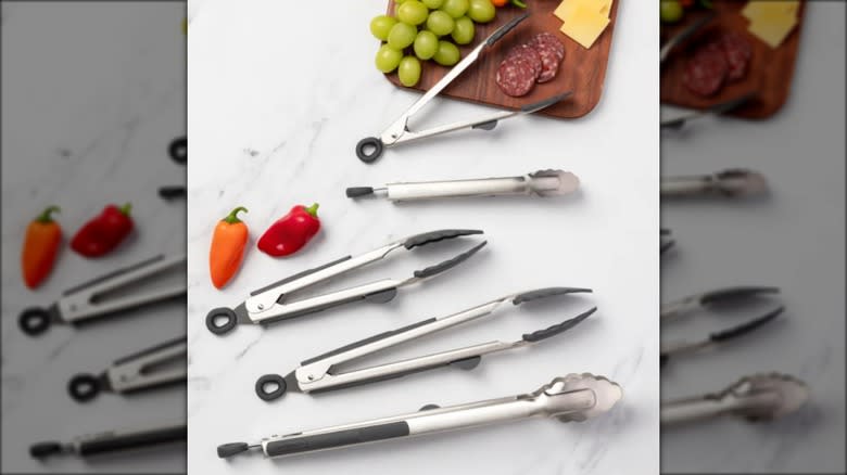 5-piece set of cooking tongs on white marble counter next to cutting board with fruit, cheese, and meat.