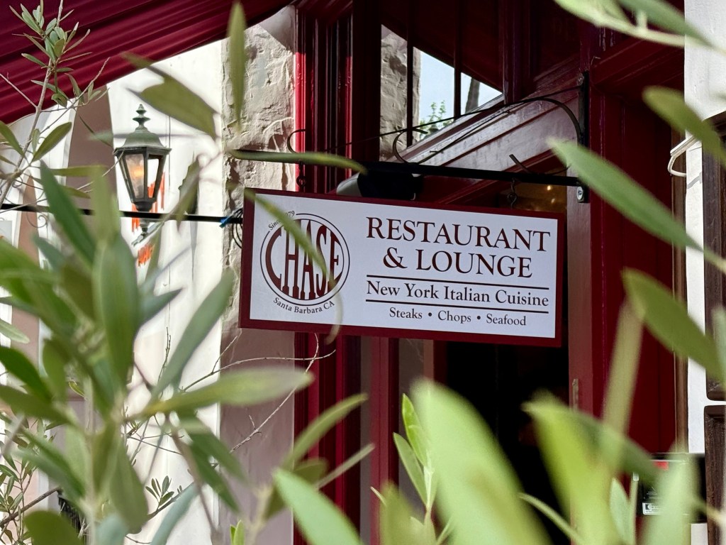 A restaurant sign for "CHASE Restaurant & Lounge" specializing in New York Italian cuisine, with foliage in the foreground.