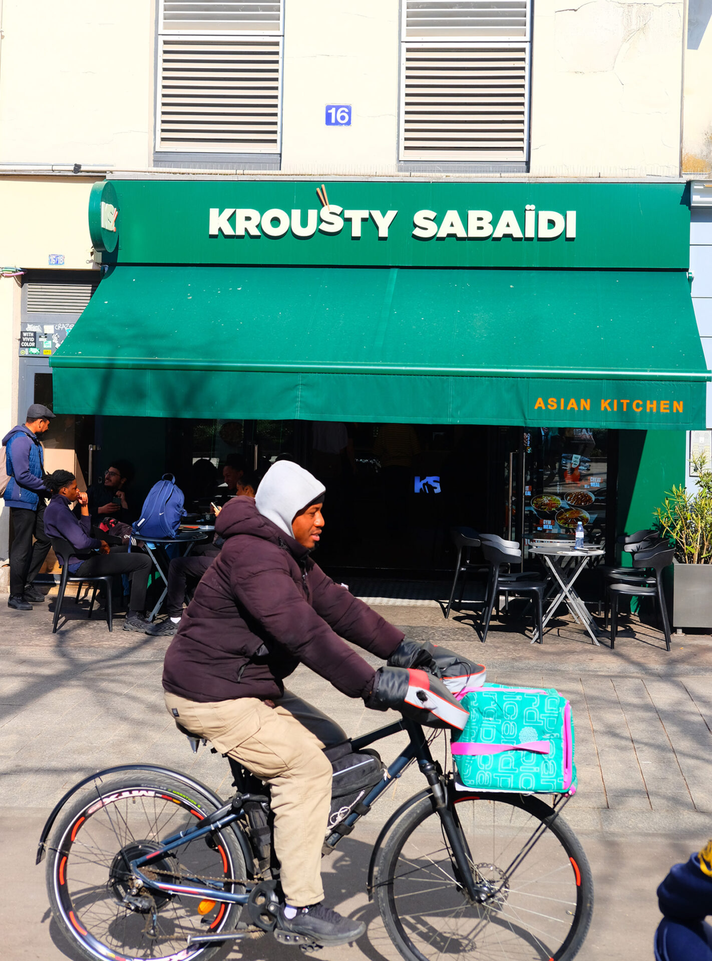 A photograph of the exterior of one of Krousty Sabaïdi's 35 restaurants, with a food delivery man on a bike cycling past
