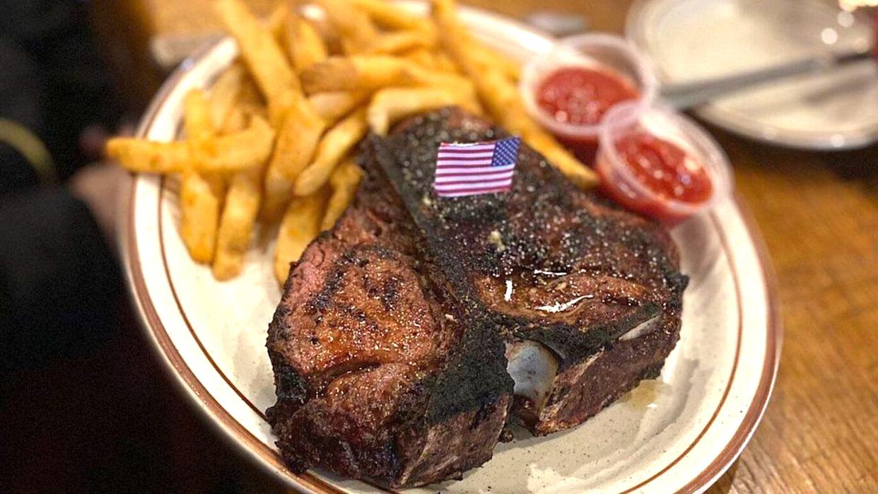 A grilled steak with a small American flag on top is served on a plate with French fries and two containers of ketchup on the side.