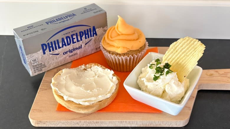 Philadelphia cream cheese on a wooden cutting board with bagel, cupcake and chip dip
