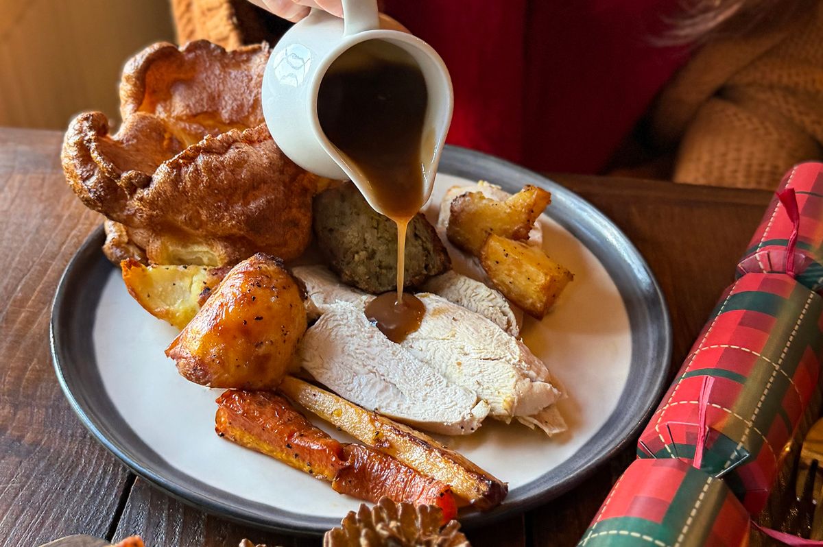 Stock photo showing close-up view of an unrecognisable, young woman in a restaurant eating a Christmas roast dinner comprising of thick slices of roast chicken, Yorkshire pudding, sage and onion bread stuffing, roasted carrots and potatoes, broccoli, cabbage, cauliflower and gravy on white plate. Also pictured are two, tartan patterned, Christmas crackers festive table decorations.