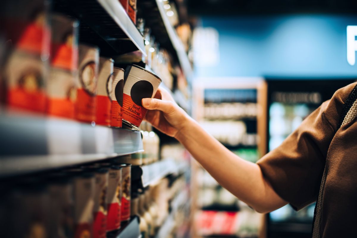 Cropped shot of young Asian woman grocery shopping in supermarket. She is picking up a tin can from the shelf and checking the nutrition label and ingredients. 