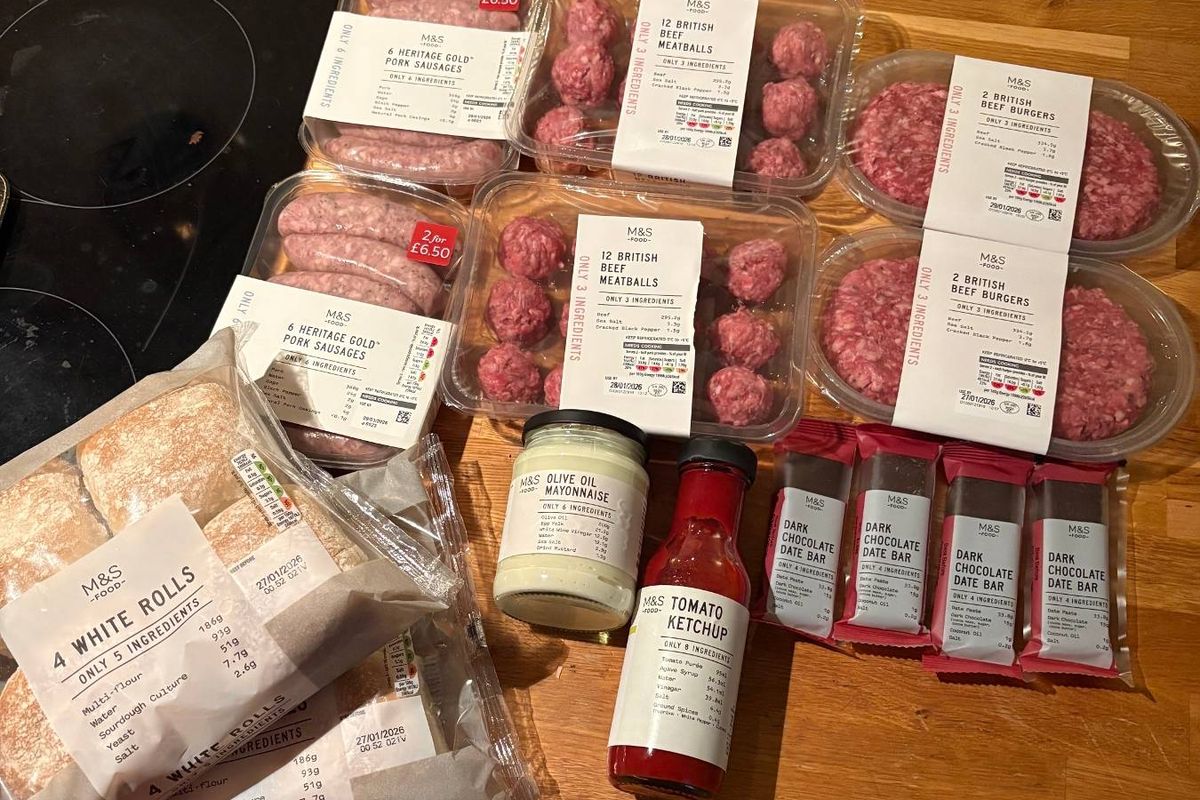 A wooden table displays an assortment of food items including red meat products, a bottle of sauce, and other packaged goods.