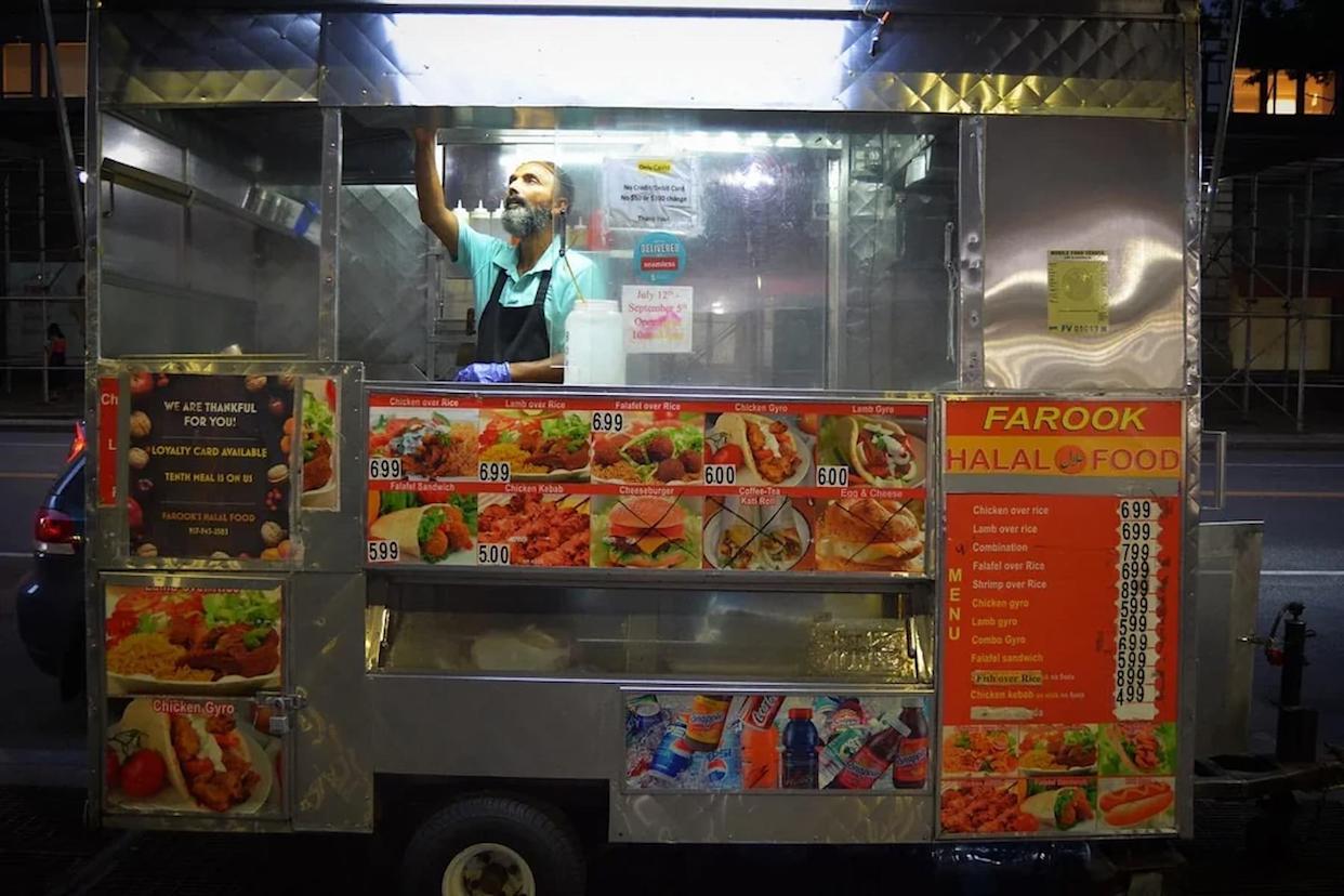 A man stands inside a brightly lit halal food cart at night, surrounded by colorful menus with photos of dishes and drinks, including gyros, kebabs, and sodas, on a city sidewalk.